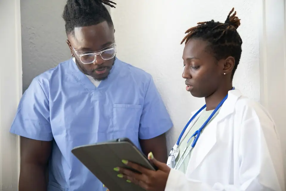 A young man in blue scrubs looking on as a physician in a white coat walks him through a chart on a tablet