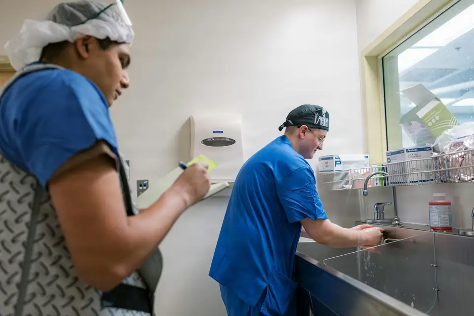 A student in scrubs taking notes on a pad while a clinician scrubs in at the sink