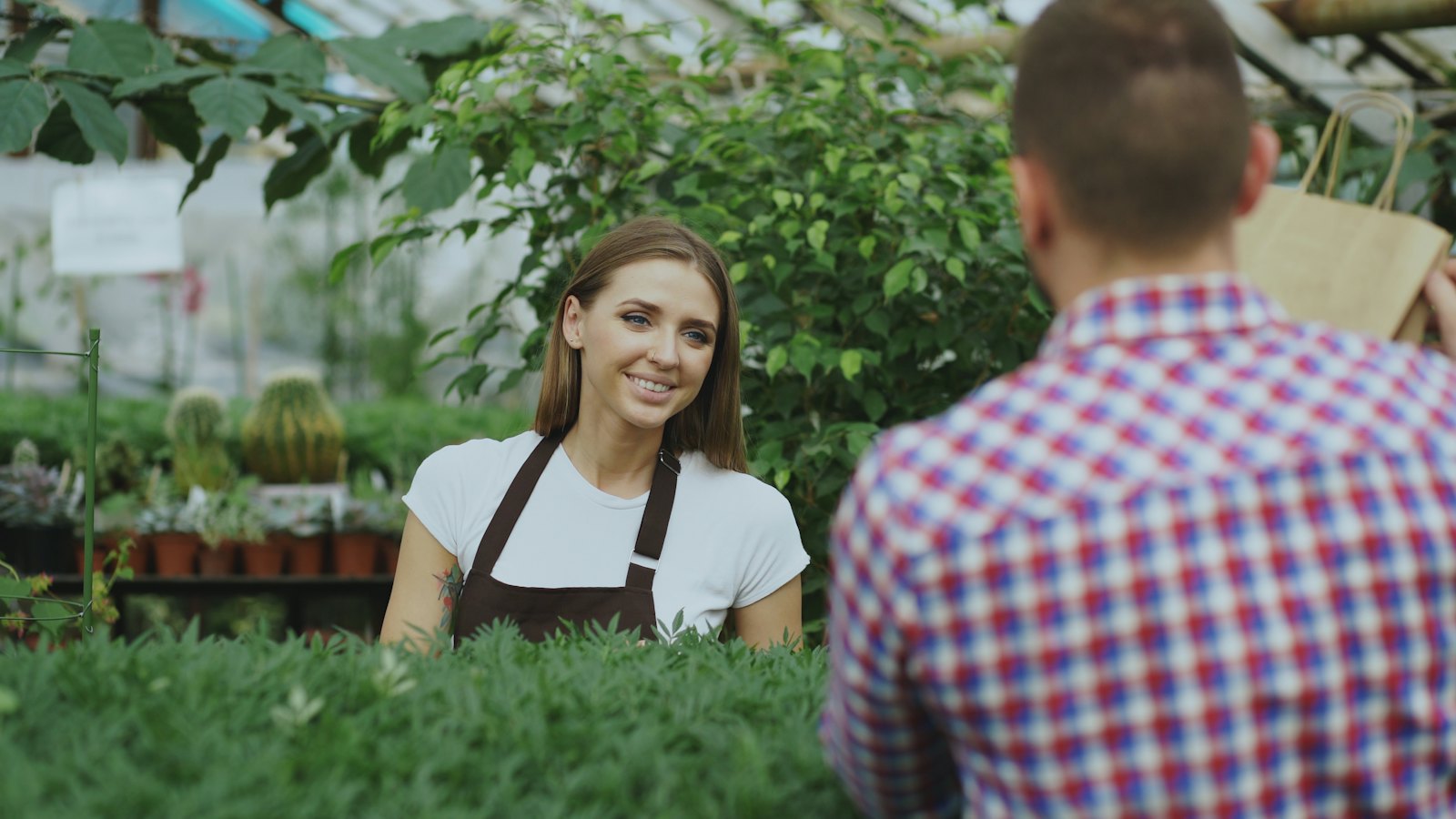 A garden center worker in an apron talking with a customer among plants in a greenhouse