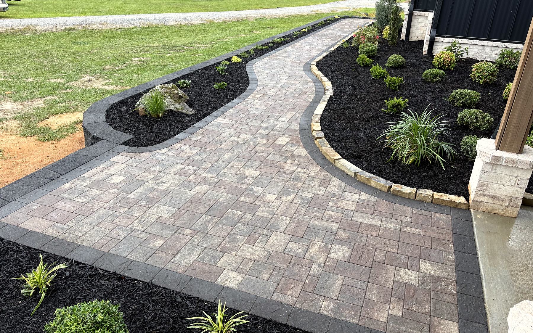 A curved paver walkway with a charcoal border and stone edging, built by Amanda's Villa between fresh flower beds at a home's front porch
