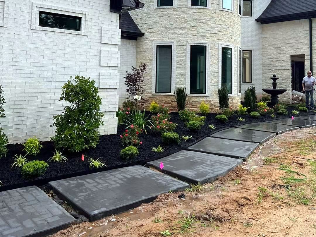 A new mulched bed of shrubs and flowers along a stone house beside a stepping-stone walk, planted by Amanda's Villa