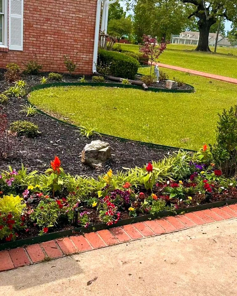 A long curved bed of annuals and shrubs with a brick border across a front lawn