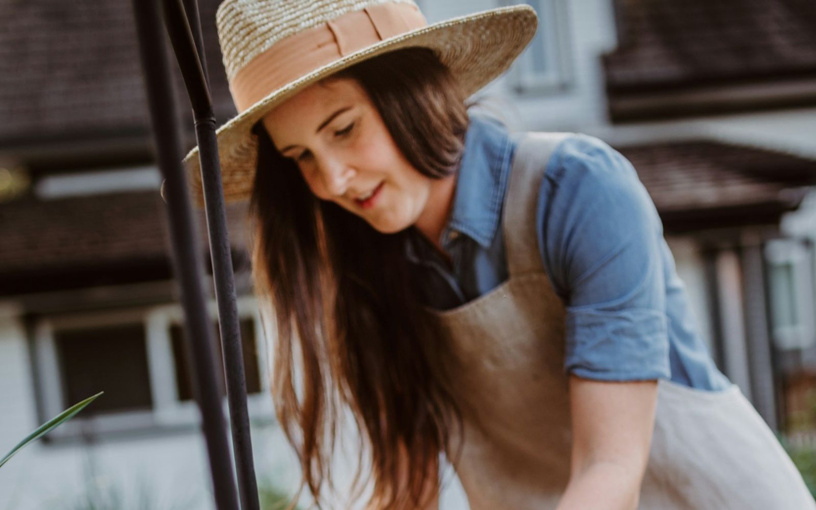 A woman in a straw hat and apron planting marigolds in a bed beside a house