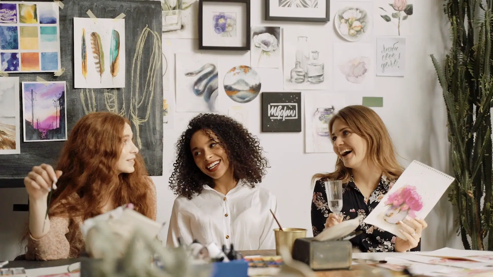 Three friends laughing at a craft table covered in watercolor supplies, one holding up her painting